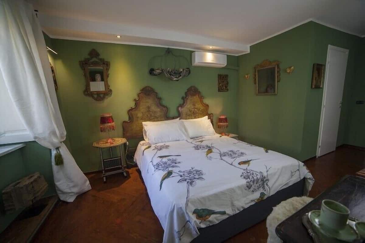 Una habitación con una cama, una mesita pequeña y una silla. Hay una ventana con cortinas, un aire acondicionado de pared y una puerta.