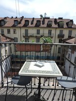 A balcony with a table and two chairs, overlooking a courtyard with buildings and a tiled roof.