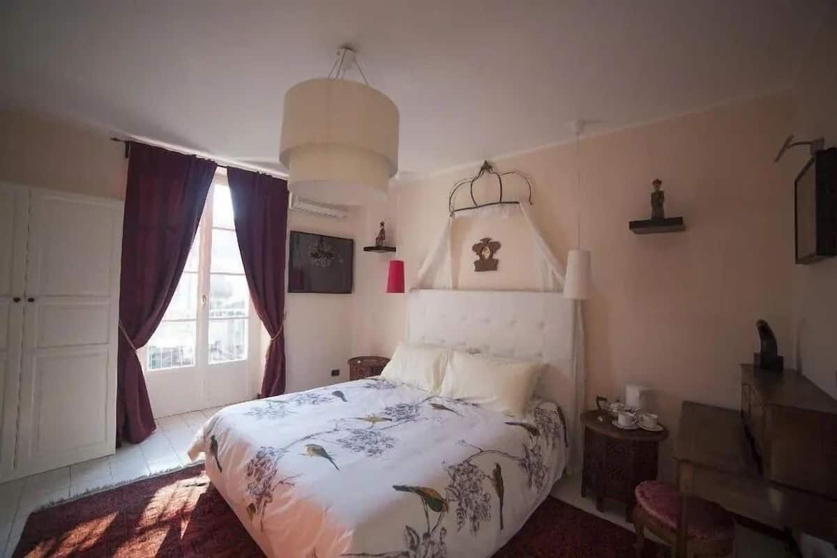 Un dormitorio con cama, mesita de noche, silla, ventana con cortinas y estante en la pared.