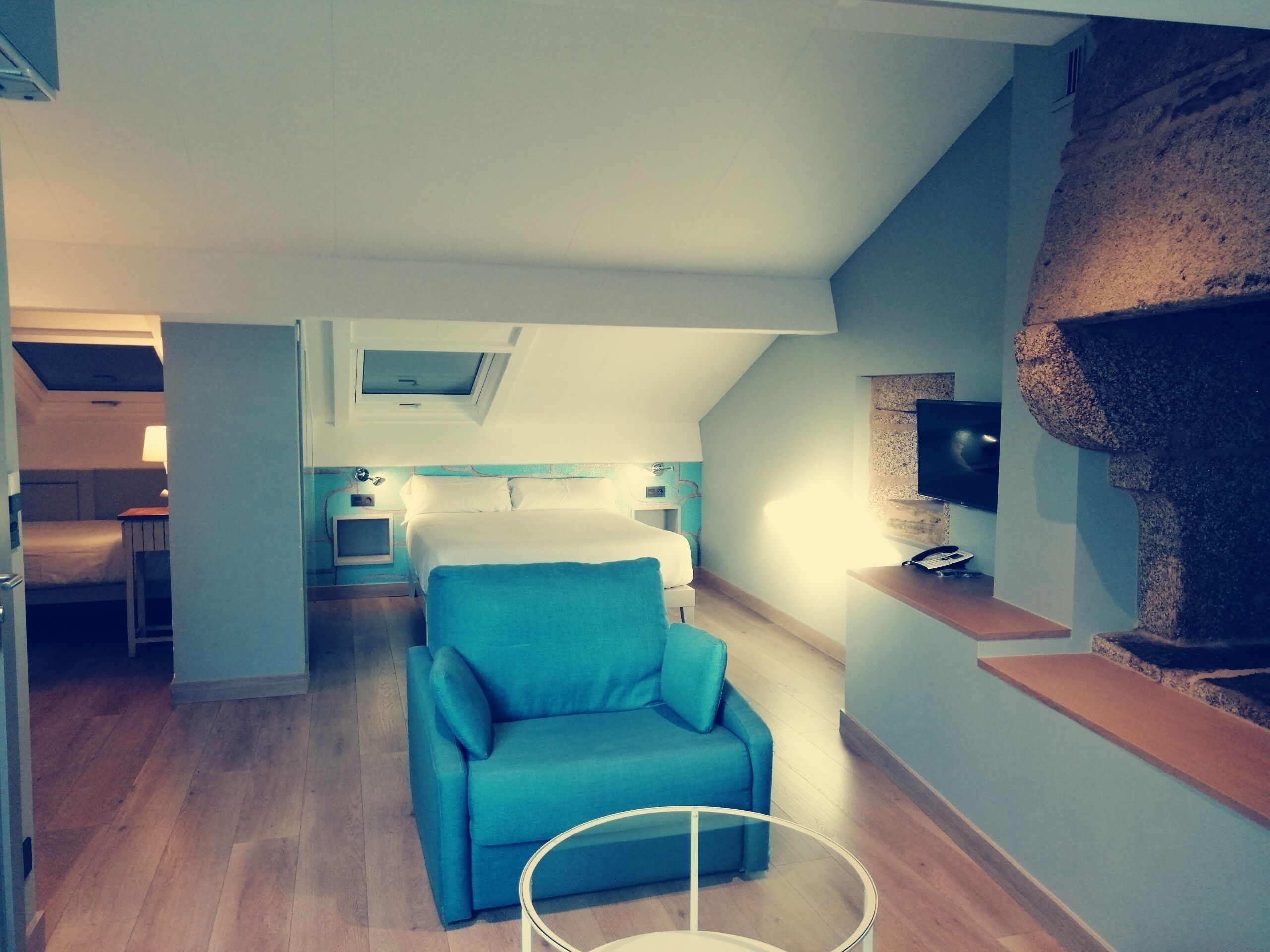 A modern hotel room with a cozy blue armchair, a small round table, a TV mounted on a stone wall, and a separate bedroom area with a bed and a desk.