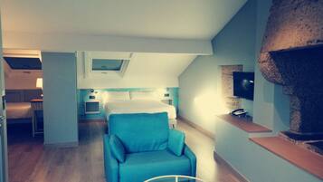 A modern hotel room with a cozy blue armchair, a small round table, a TV mounted on a stone wall, and a separate bedroom area with a bed and a desk.