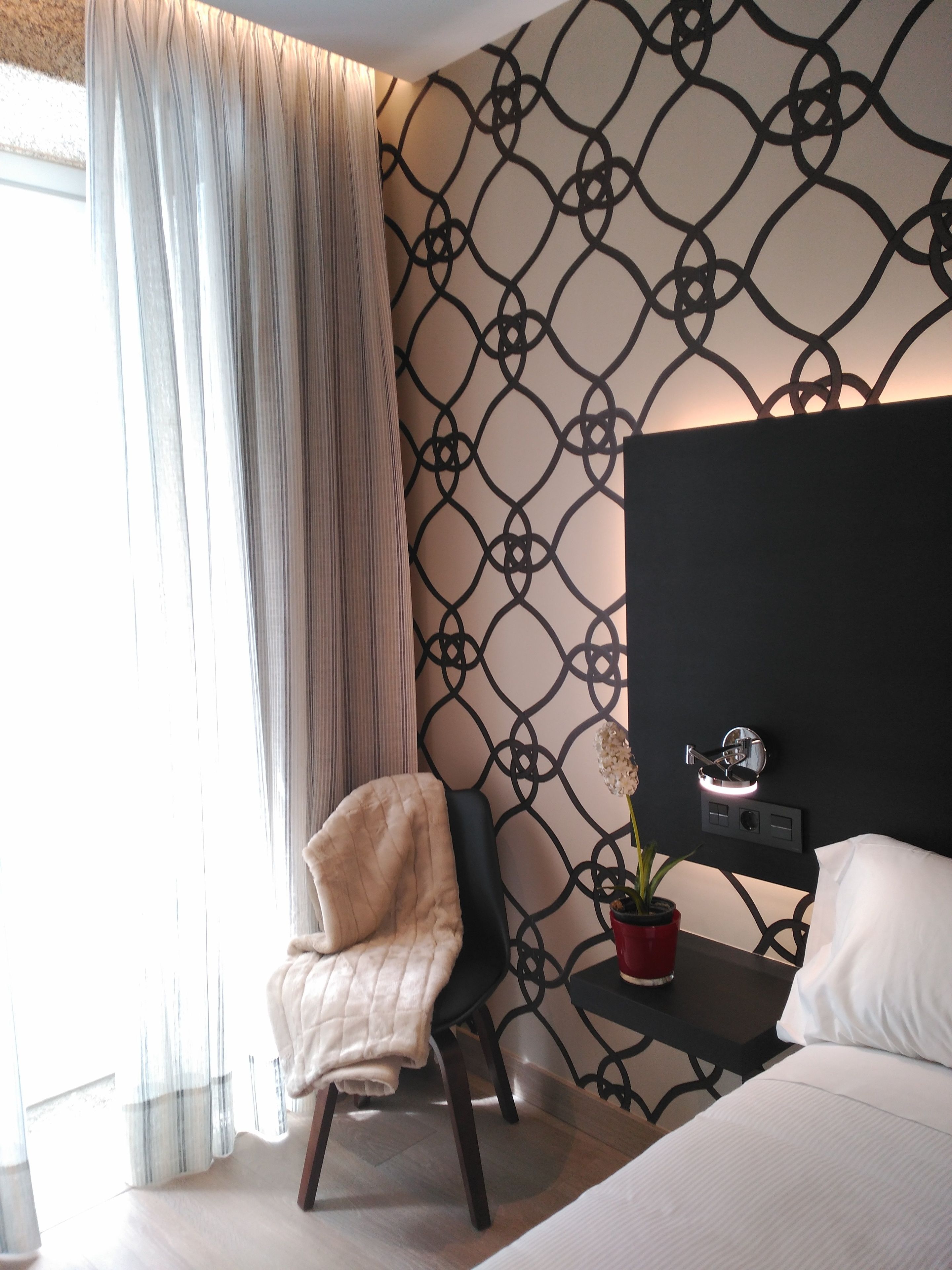 A modern hotel room with a patterned wall, a large bed, a bedside table with a potted plant, and a chair with a throw blanket.