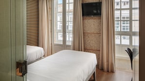 Hotellihuone, jossa on kaksi sänkyä, seinälle asennettu televisio ja suuri ikkuna, josta avautuu näkymä ympäröiviin rakennuksiin.
