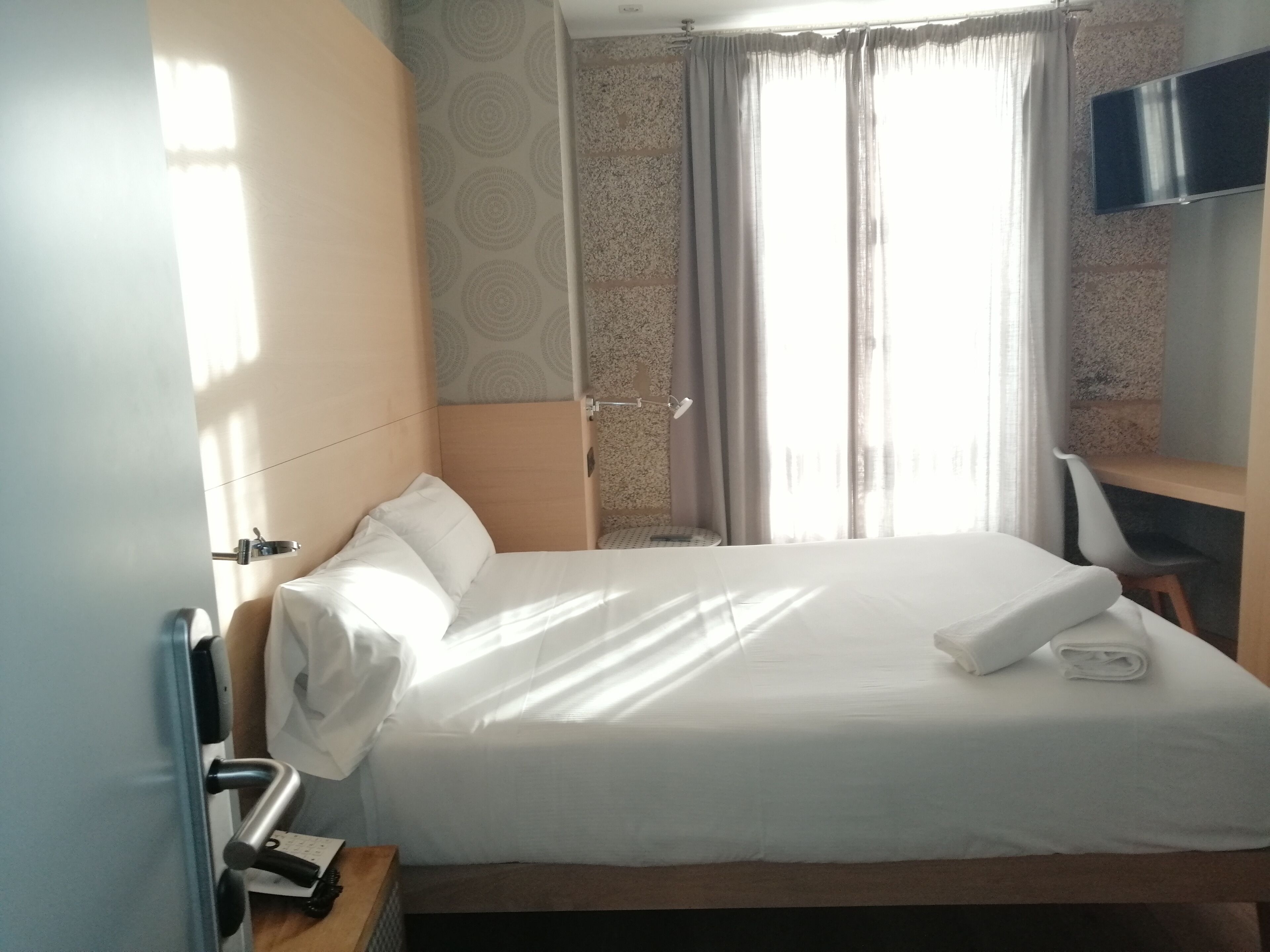 A hotel room with a bed, a desk, a chair, a TV, and a window with curtains.