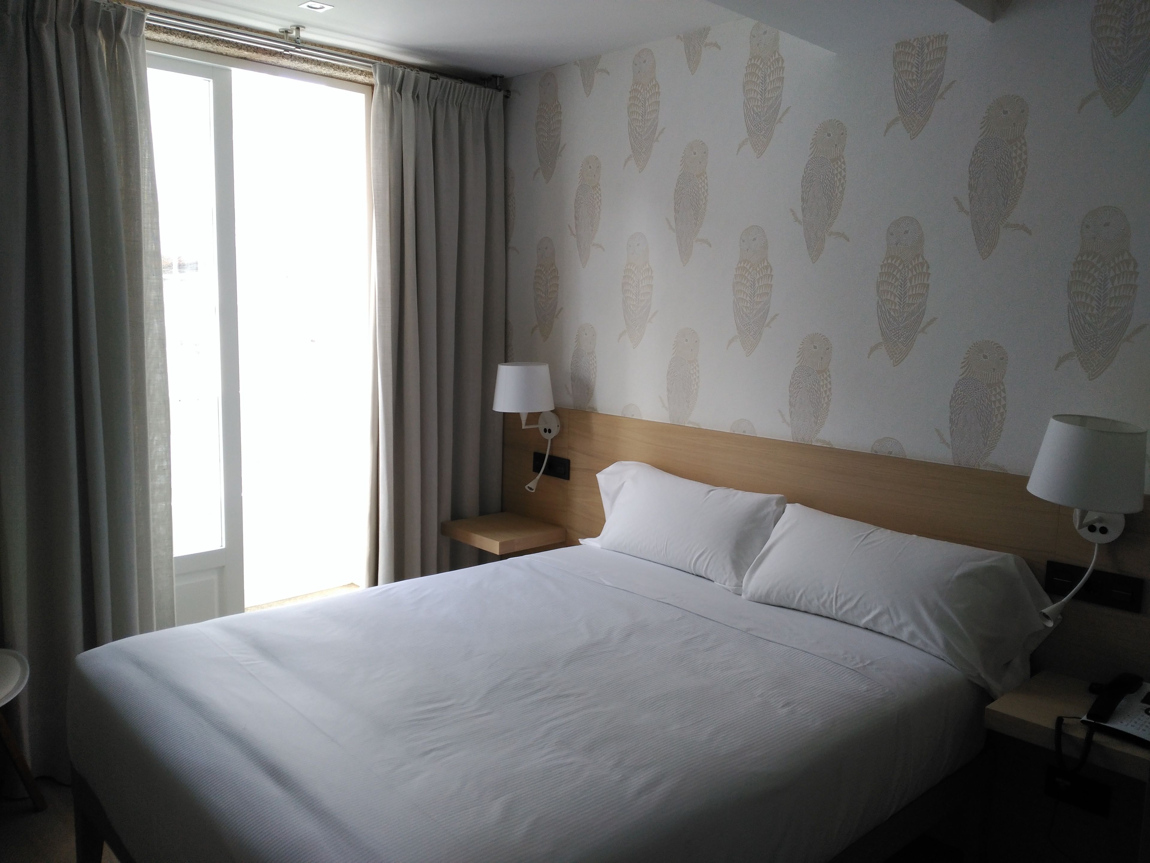 Une chambre d’hôtel avec un grand lit, deux lampes de chevet, une tête de lit en bois et une fenêtre avec des rideaux.