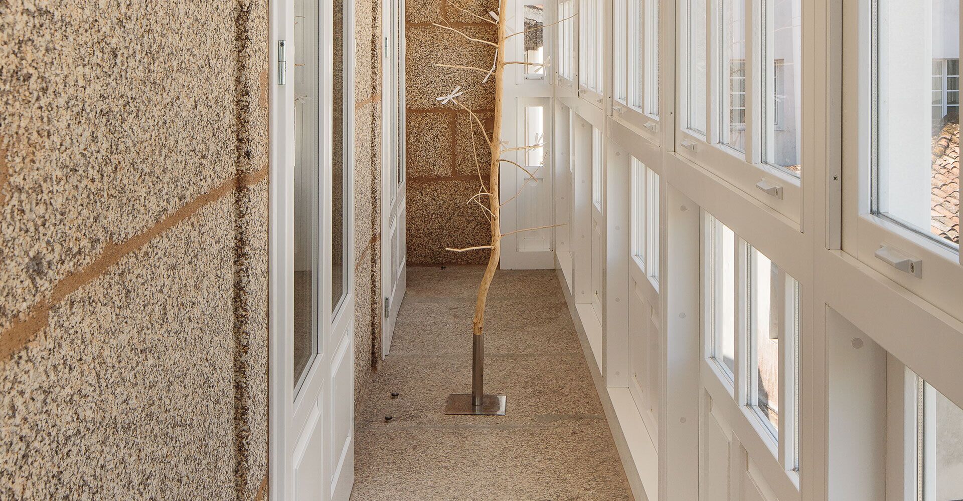 Een gang met witte wanden, een gestructureerde vloer en een houten constructie in het midden.