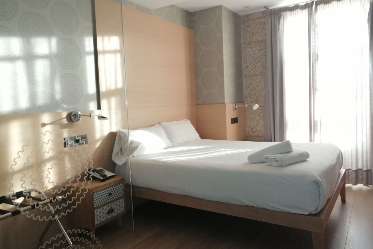 Una habitación de hotel moderna con una cama de madera, una mesita de noche y un amplio ventanal con cortinas.