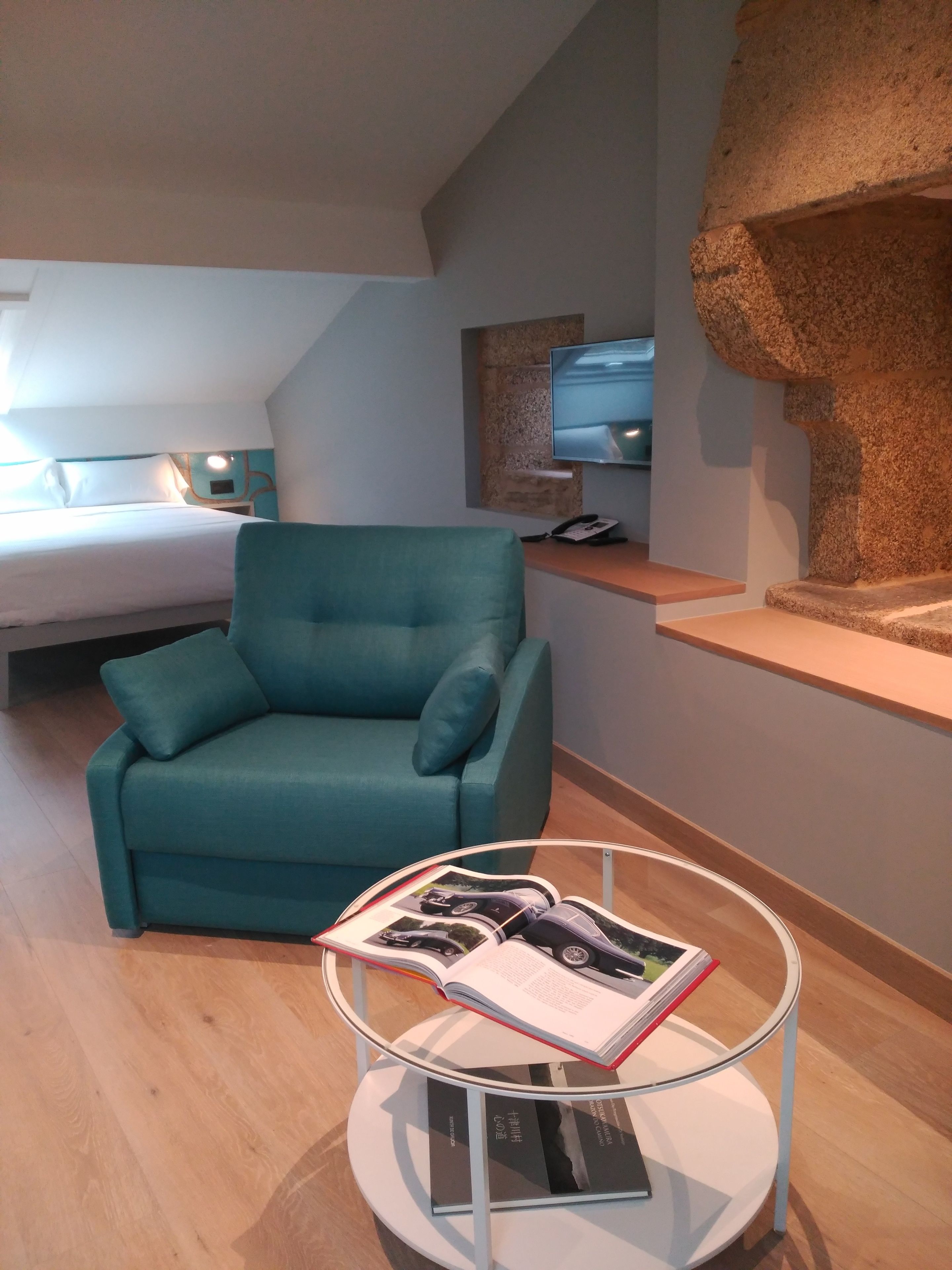 A modern hotel room with a teal sofa, a round glass table with books, a bed with white linens, and a stone accent wall.