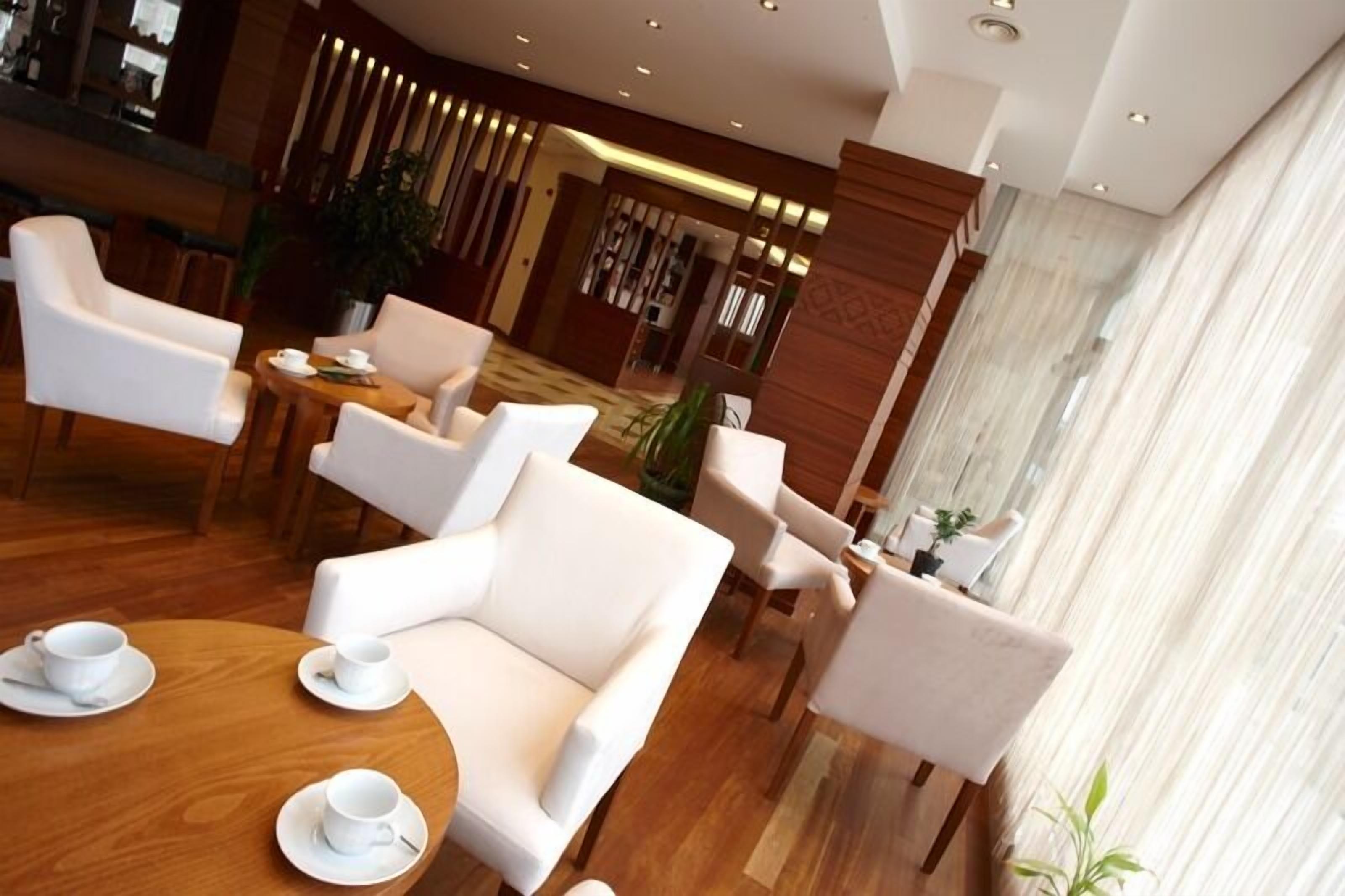 A modern hotel lobby with white leather sofas, wooden tables, and a bar area in the background.
