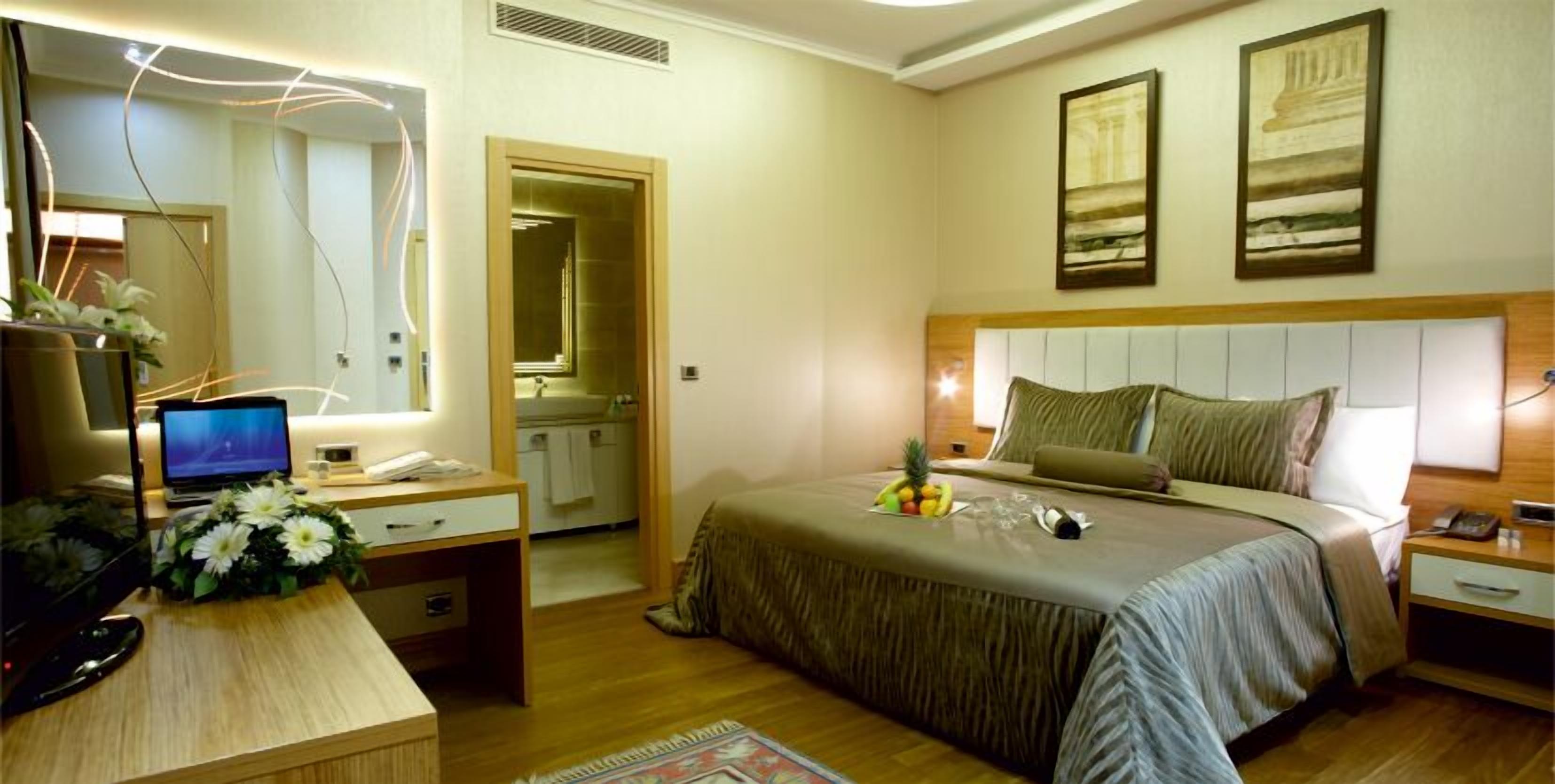 A hotel room with a bed, bedside tables, a desk with a computer, a mirror, and artwork on the walls.