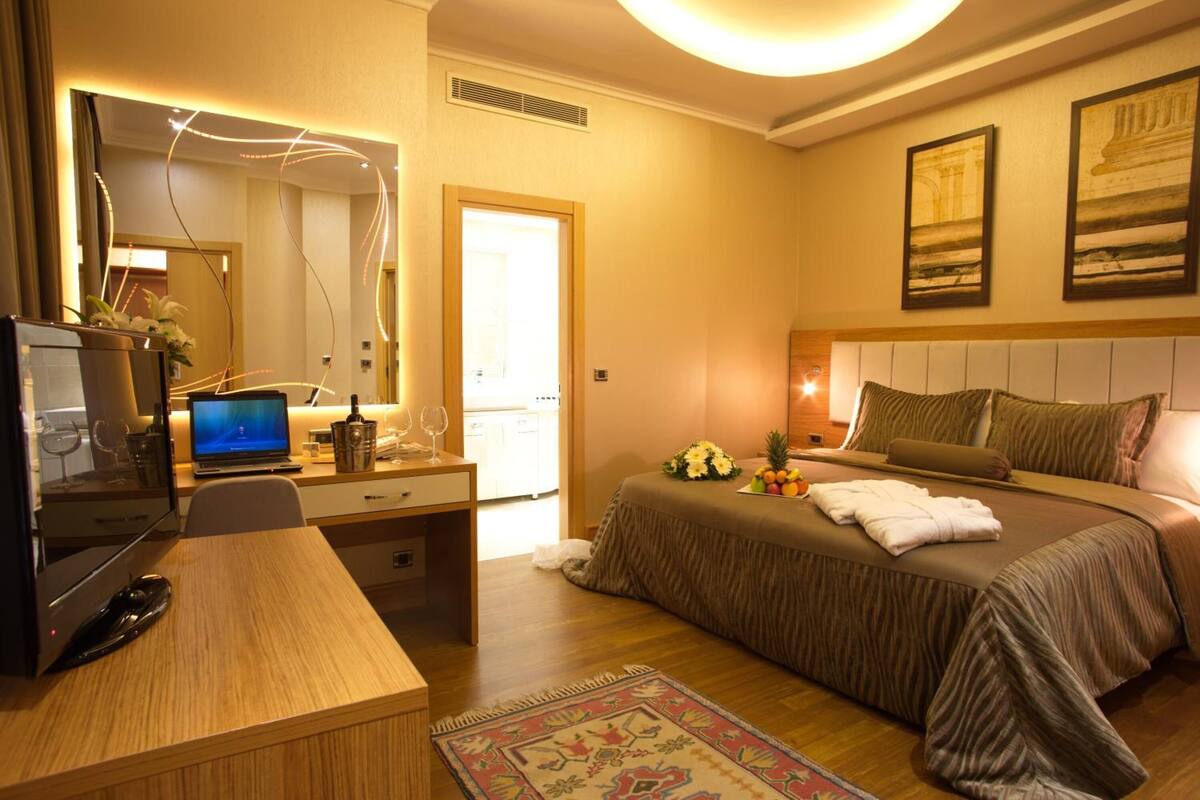 A hotel room with a bed, a desk with a laptop, a mirror, and a small table with a vase of flowers.
