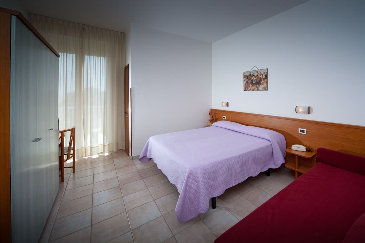 Habitación de hotel con cama, sofá rojo, cabecero de madera, mesita, silla y un cuadro en la pared.