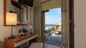 Una habitación con un escritorio de madera, una silla, un televisor, un espejo y un balcón con vistas al mar y edificios.