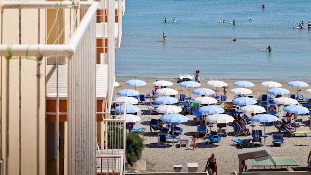 Una playa con sombrillas y sillas reclinables, un cielo azul despejado y un edificio con balcones que dan al mar.