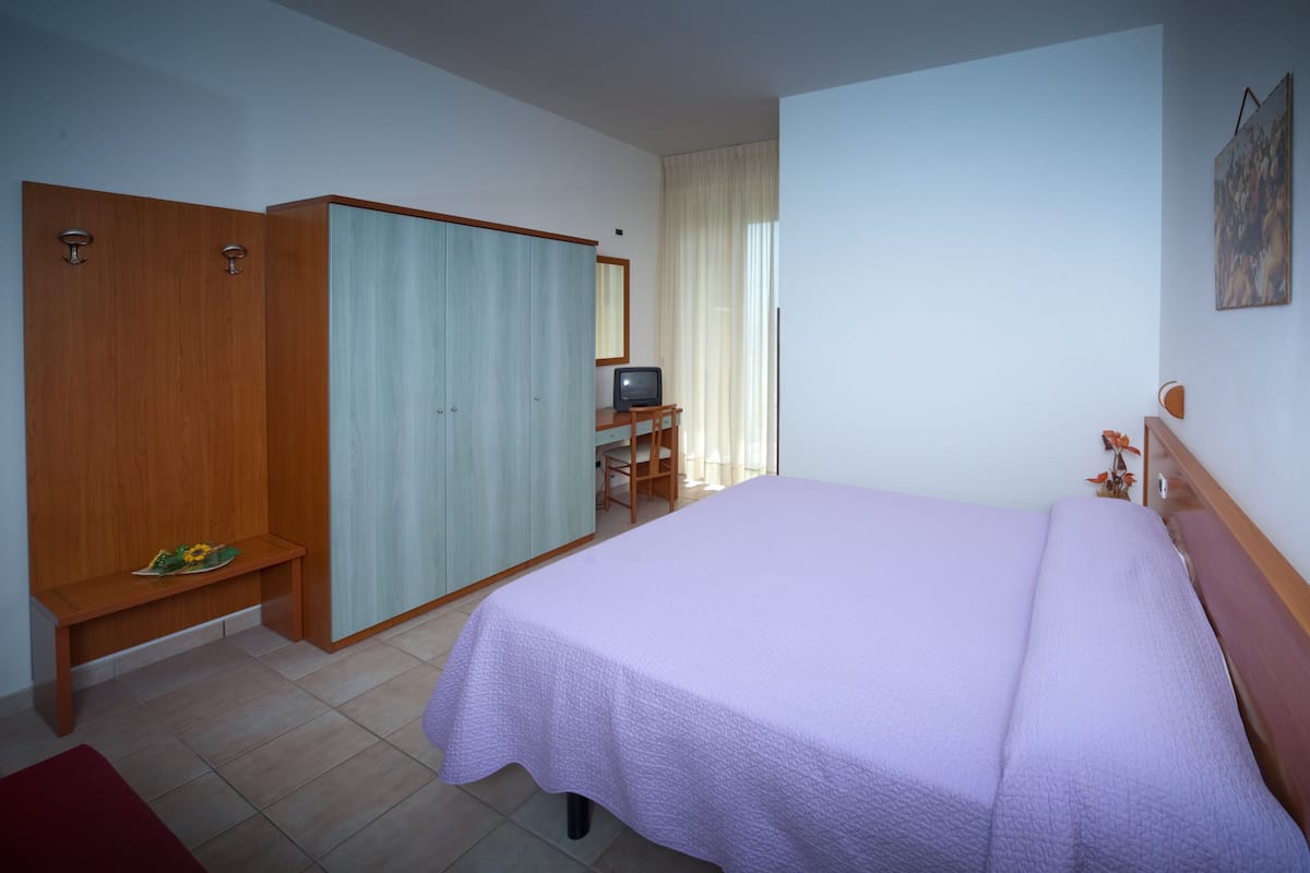 Un dormitorio con una cama, un armario de madera, un escritorio con televisor y una ventana con cortinas.