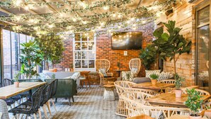 Een terras buiten met houten tafels en stoelen, omgeven door potplanten en lichtslingers.