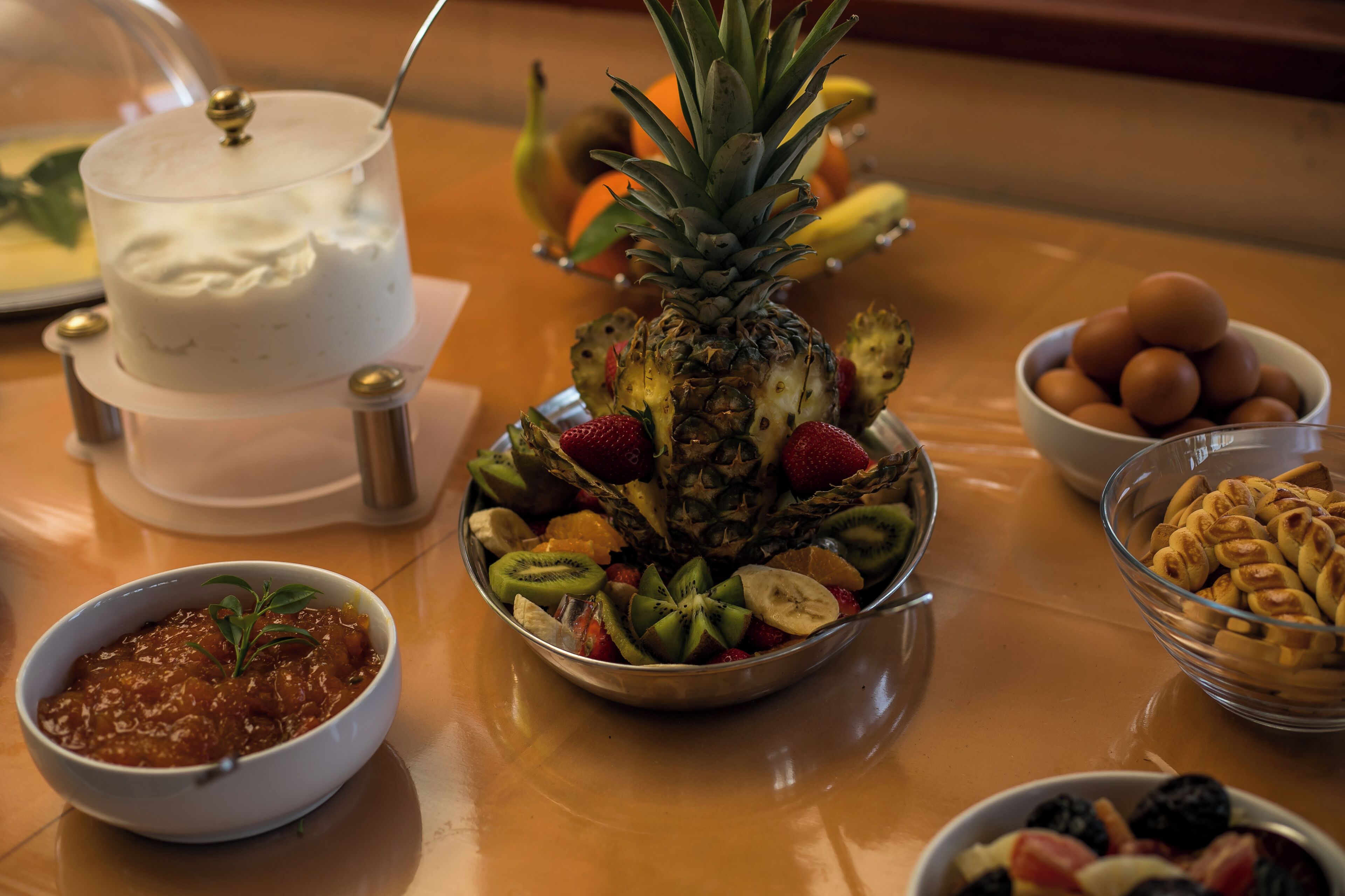 Eine Obstplatte mit Ananas, Erdbeeren, Kiwi und anderem Obst, eine Schale mit Eiern sowie ein Dessert mit Glaskuppel.