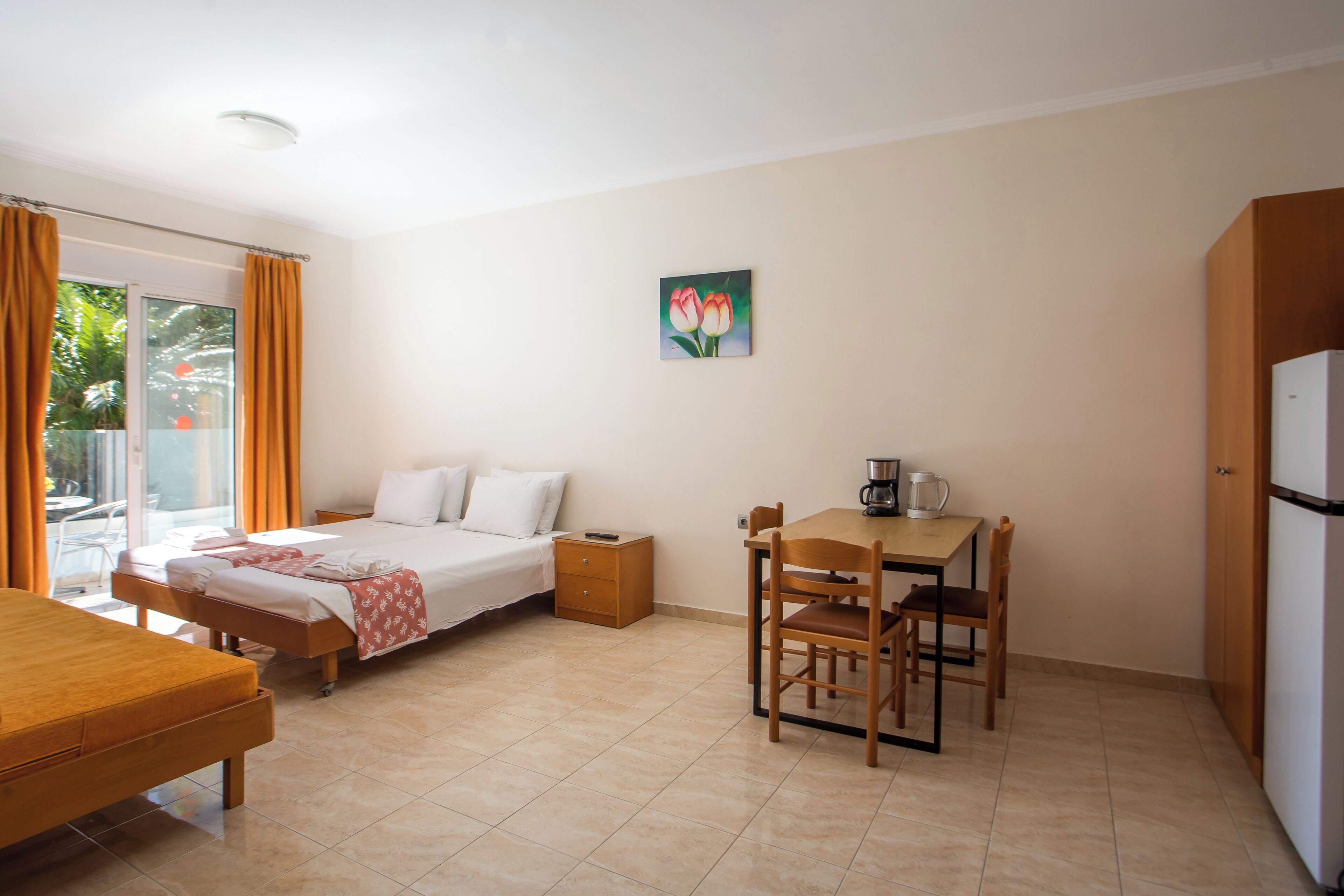 A hotel room with a bed, a desk with a chair, a small table, a refrigerator, and a painting on the wall.