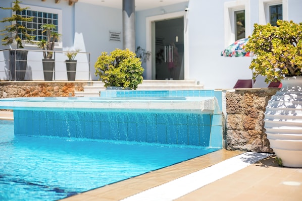 A swimming pool with clear blue water, surrounded by potted plants and a stone wall. - Perla Marina (Rhodes)