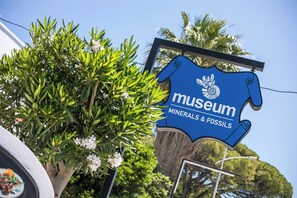 A museum sign with a blue background and white text, surrounded by greenery and a clear sky. - Perla Marina (Rhodes)