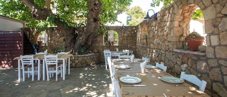 An outdoor dining area with stone walls, white furniture, and a large tree providing shade.