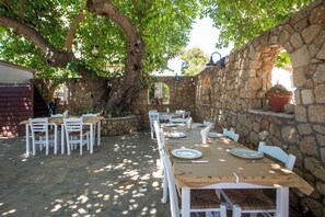 An outdoor dining area with stone walls, white furniture, and a large tree providing shade. - Perla Marina (Rhodes)