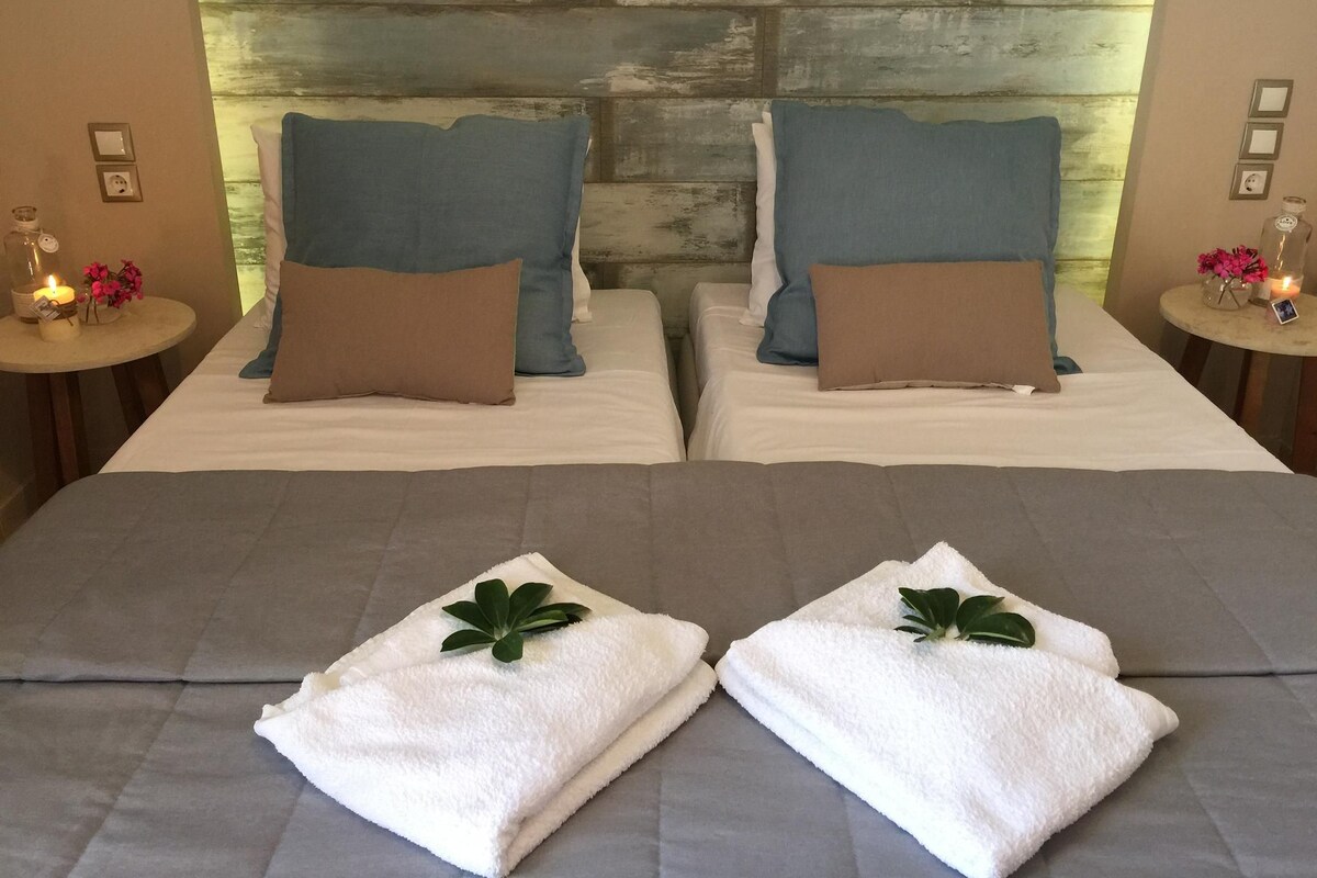 Deux lits avec des oreillers bleus et bruns, une tête de lit en bois, des tables de chevet avec des bougies et des serviettes pliées ornées de feuilles vertes.