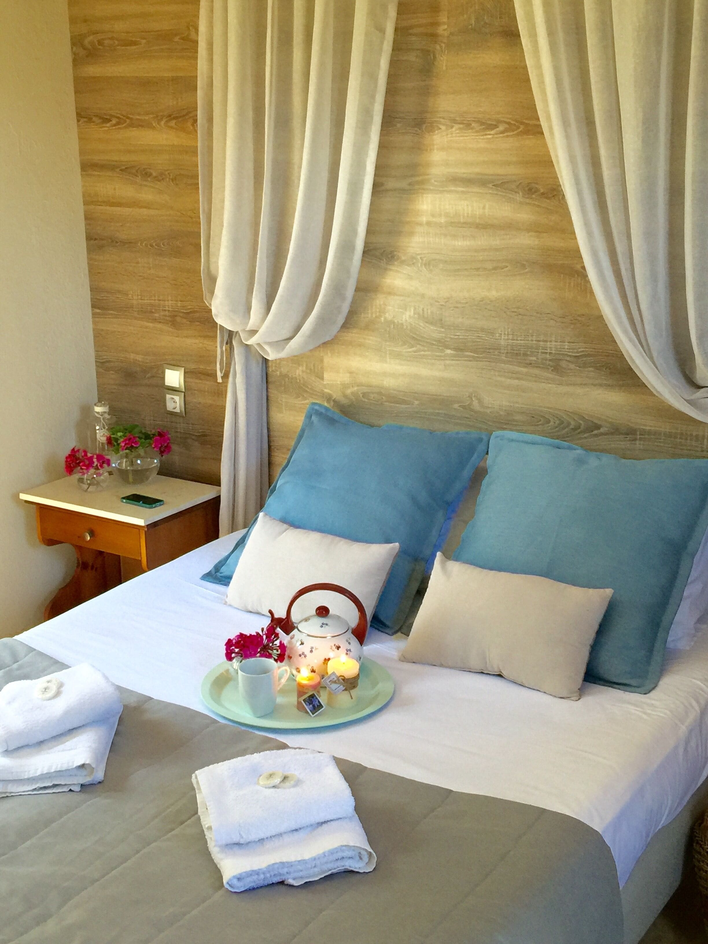 A neatly made bed with blue and white pillows, a bedside table with a phone and a glass container with flowers, and a wooden headboard with draped curtains.