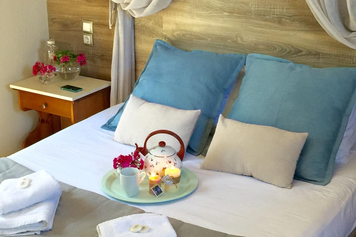 A neatly made bed with blue and white pillows, a bedside table with a phone and a glass container with flowers, and a wooden headboard with draped curtains.