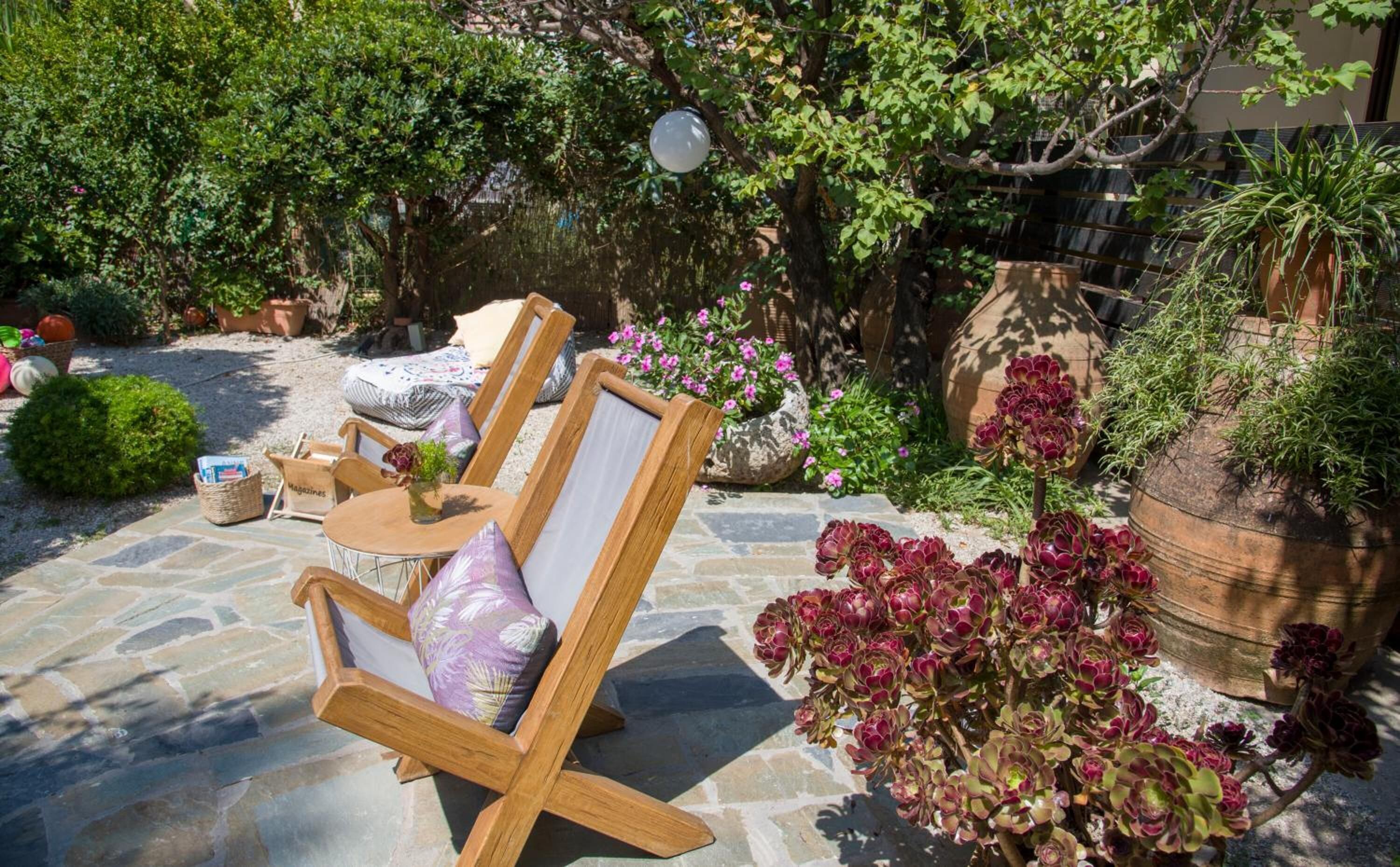A garden patio with stone flooring, two wooden chairs, a small table, and various potted plants.