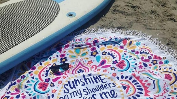 Ein buntes Strandtuch mit einem motivierenden Spruch und einem aufblasbaren Schlauchboot in Blau und Weiß auf einem Sandstrand.