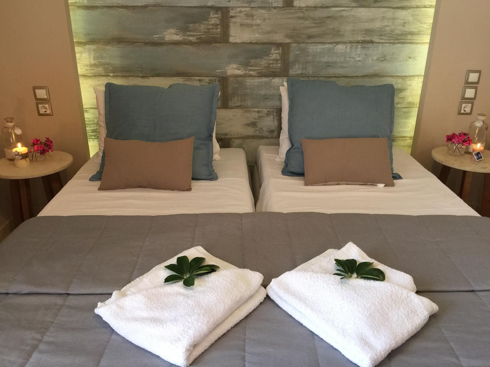 Two beds with blue and brown pillows, a wooden headboard, bedside tables with candles, and folded towels with green leaves.