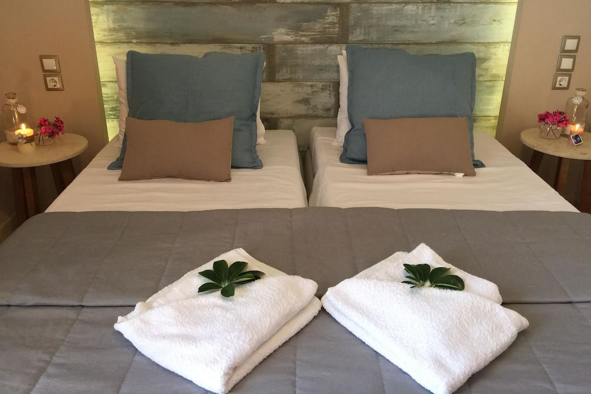 Two beds with blue and brown pillows, a wooden headboard, bedside tables with candles, and folded towels with green leaves.