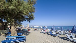 Ein Strand mit Reihen von Liegestühlen und Sonnenschirmen, ein Baum und ein klarer, blauer Himmel.