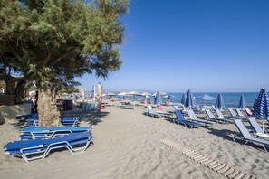Une plage bordée de rangées de transats et de parasols, un arbre et un ciel bleu azur.