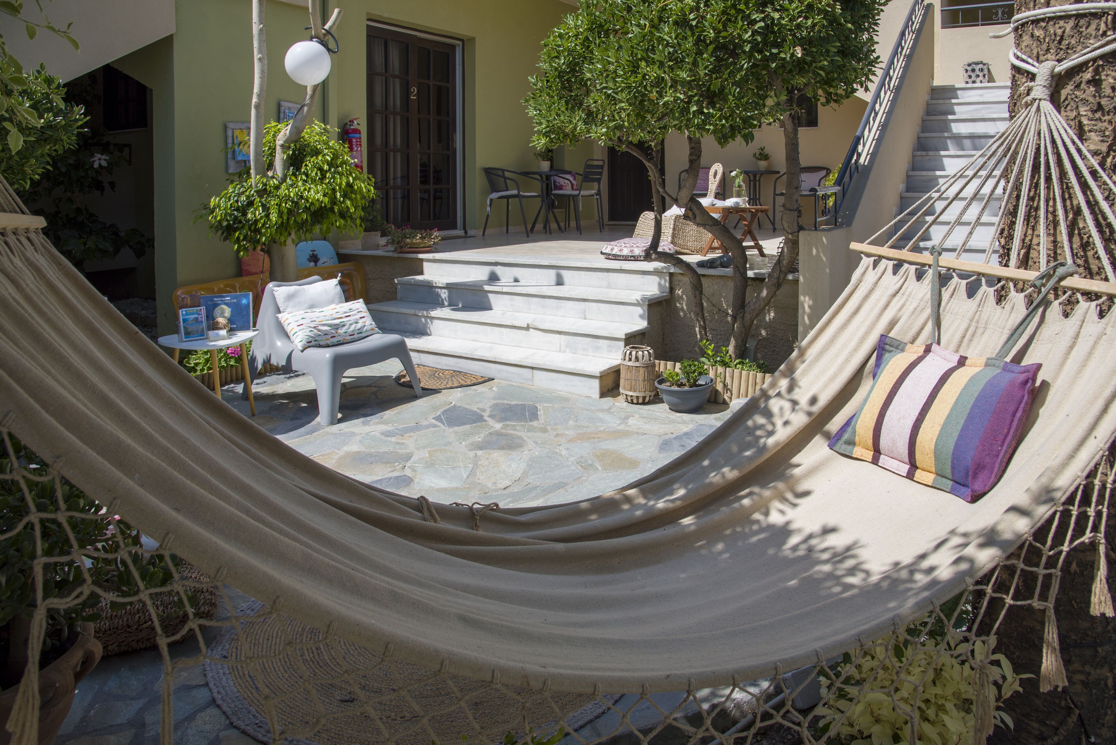 A hammock in a sunlit outdoor area with a stone patio, a staircase, and a dining table with chairs.