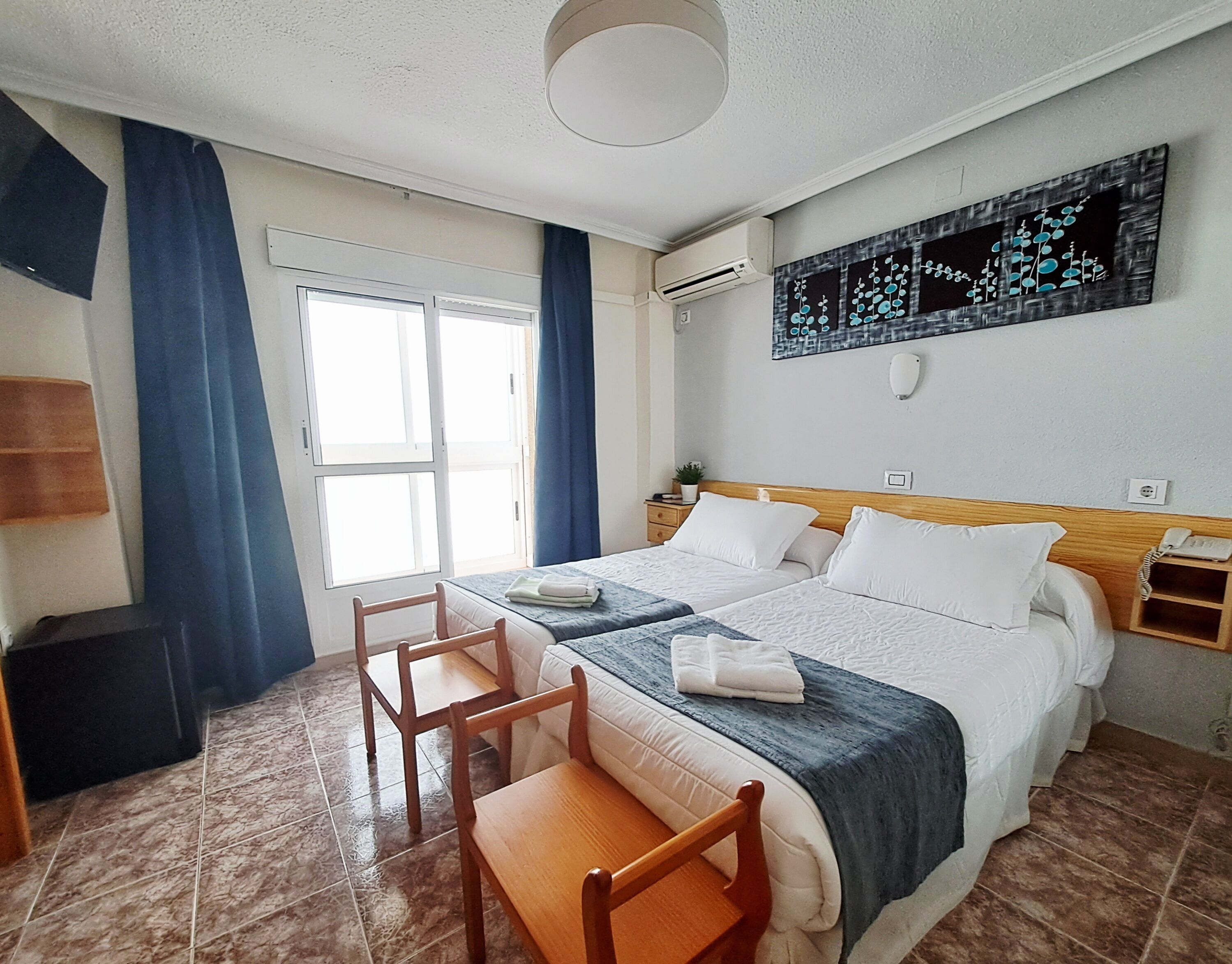 Habitación de hotel con cama, dos mesitas de noche, una silla, ventana con cortinas, equipo de aire acondicionado y una obra de arte colgada en la pared.