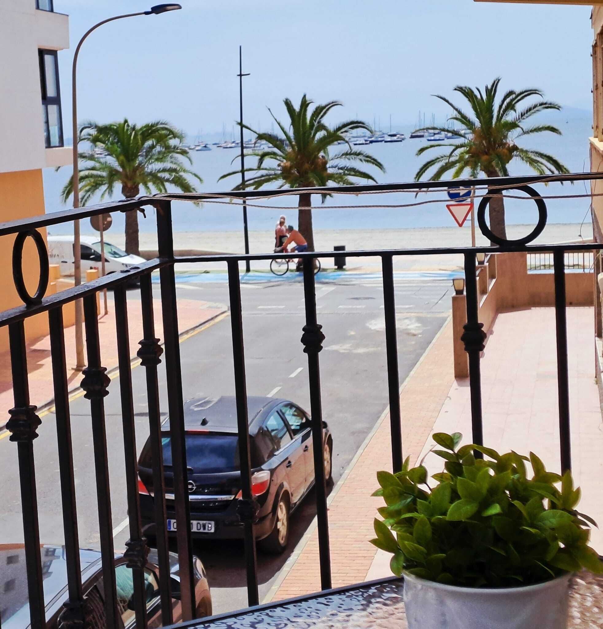 A balcony with a view of a street, parked cars, and a distant beach with boats.