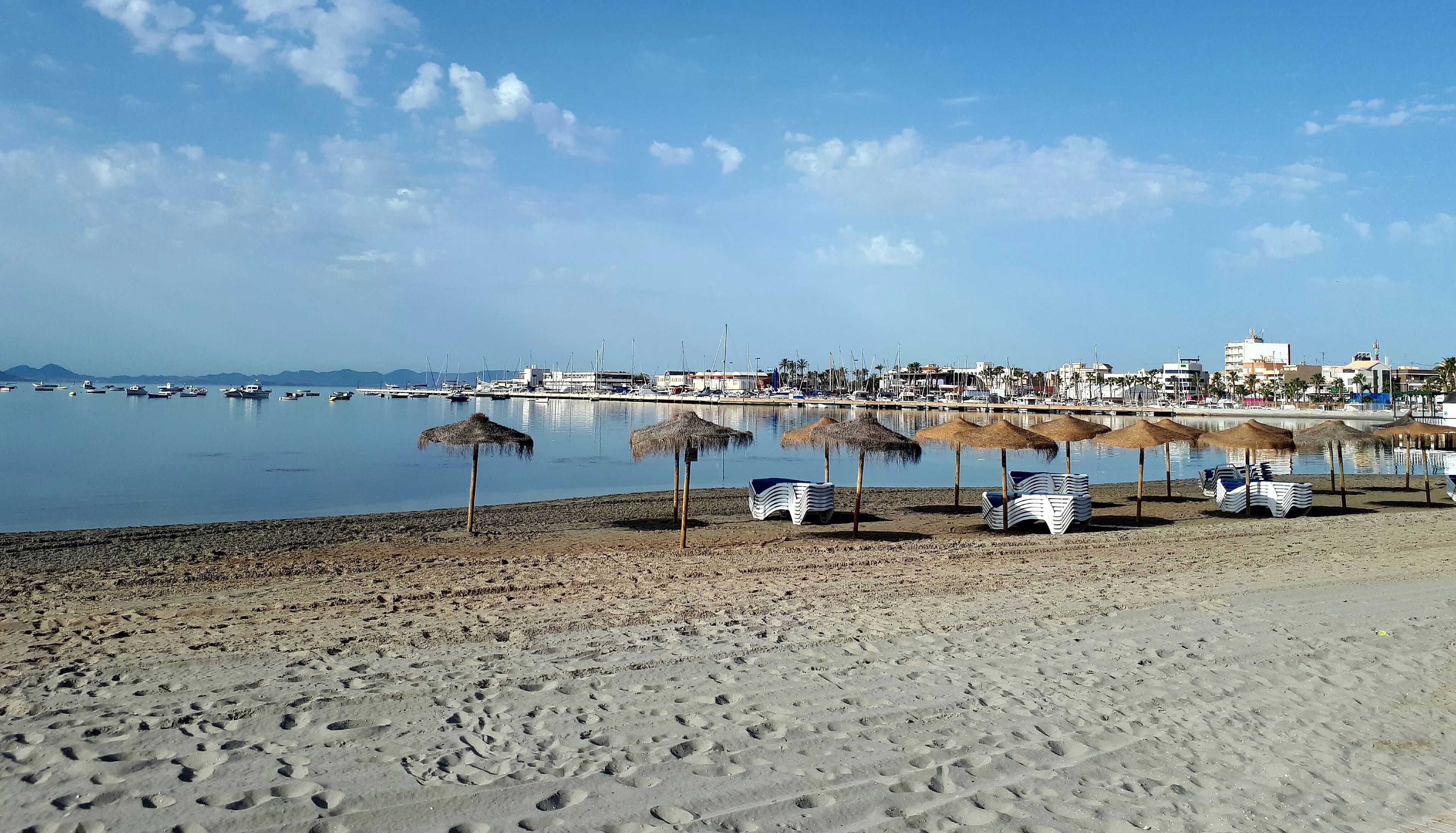 A beach with umbrellas and lounge chairs, a marina with boats, and buildings in the background.