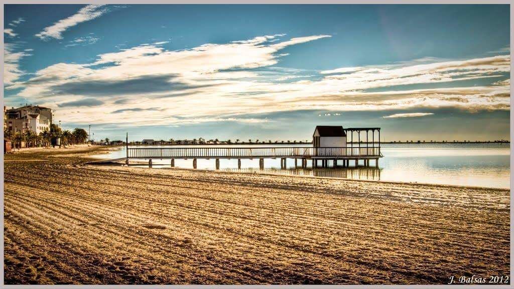 Une plage avec une jetée en bois qui s’avance dans une eau calme, un petit bâtiment sur la jetée et une côte lointaine parsemée de bâtiments.