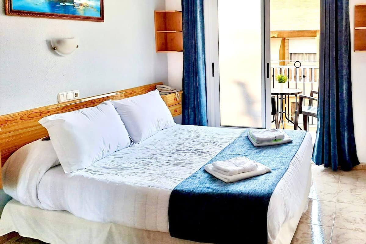 Ein Hotelzimmer mit Bett, Nachttisch, Wandleuchte, einem Fenster mit Vorhängen und Blick auf den Außenbalkon.