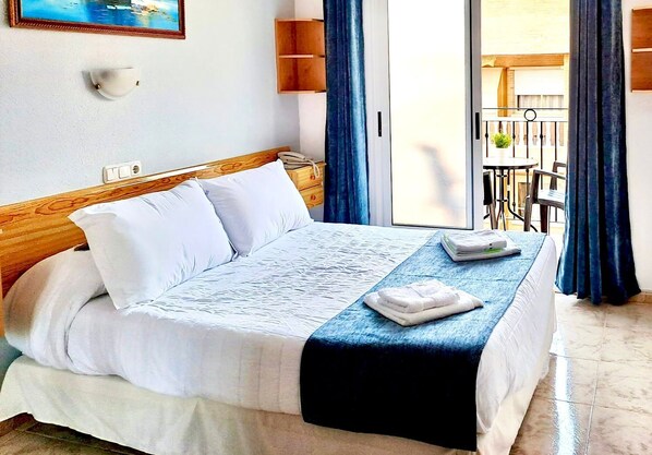 Habitación de hotel con cama, mesita de noche, lámpara de pared, ventana con cortinas y vista al balcón exterior.