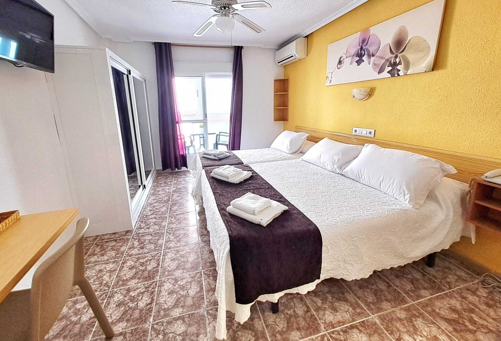 Camera d'albergo con un letto, una scrivania, una sedia, un televisore, una ventola a soffitto e una finestra con tende.