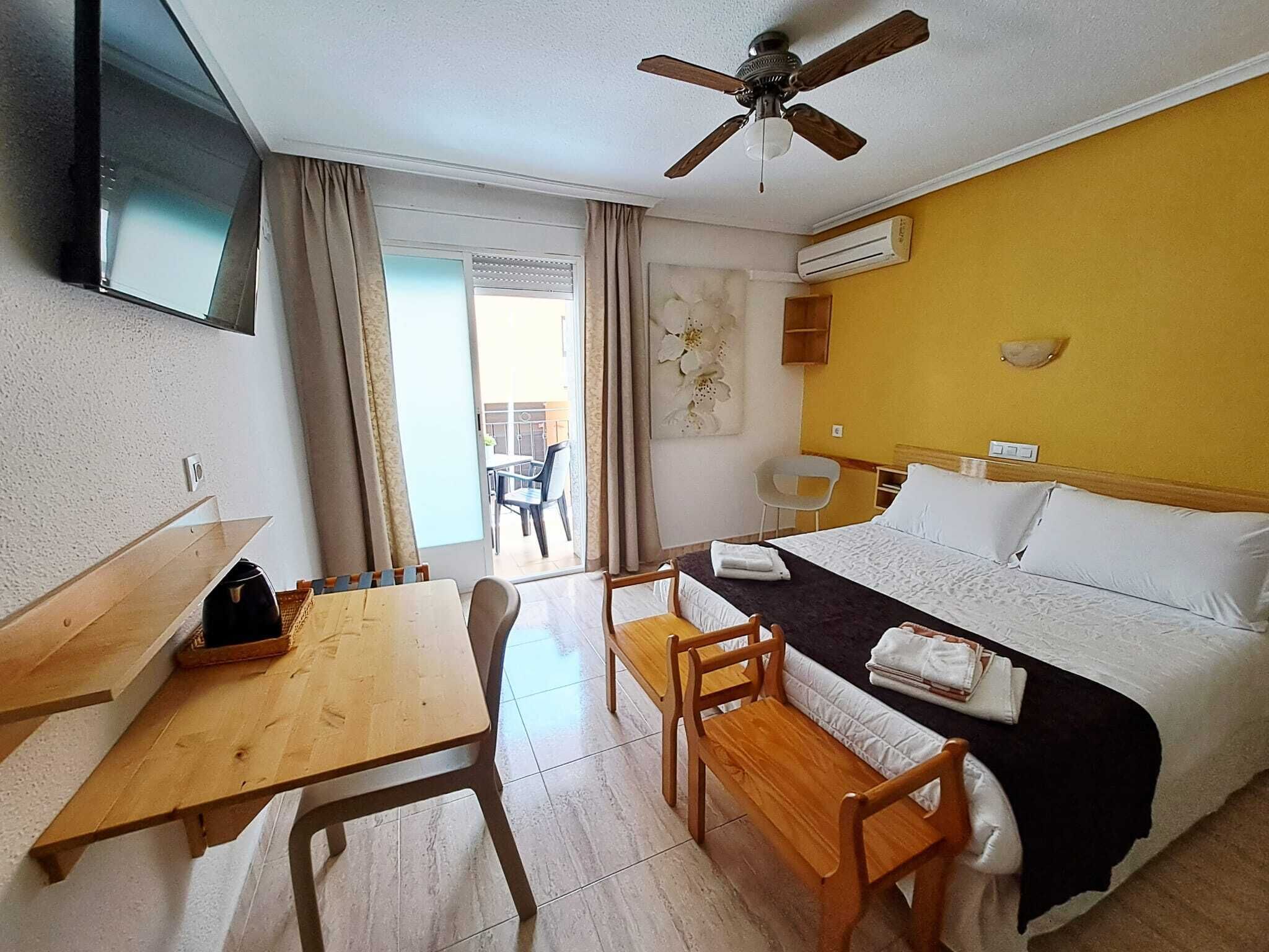 A hotel room with a bed, a desk, a chair, a table, a ceiling fan, and an air conditioning unit.