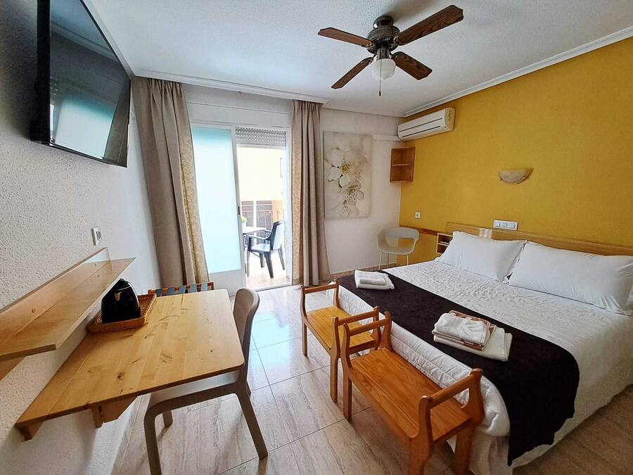 A hotel room with a bed, a desk, a chair, a table, a ceiling fan, and an air conditioning unit.