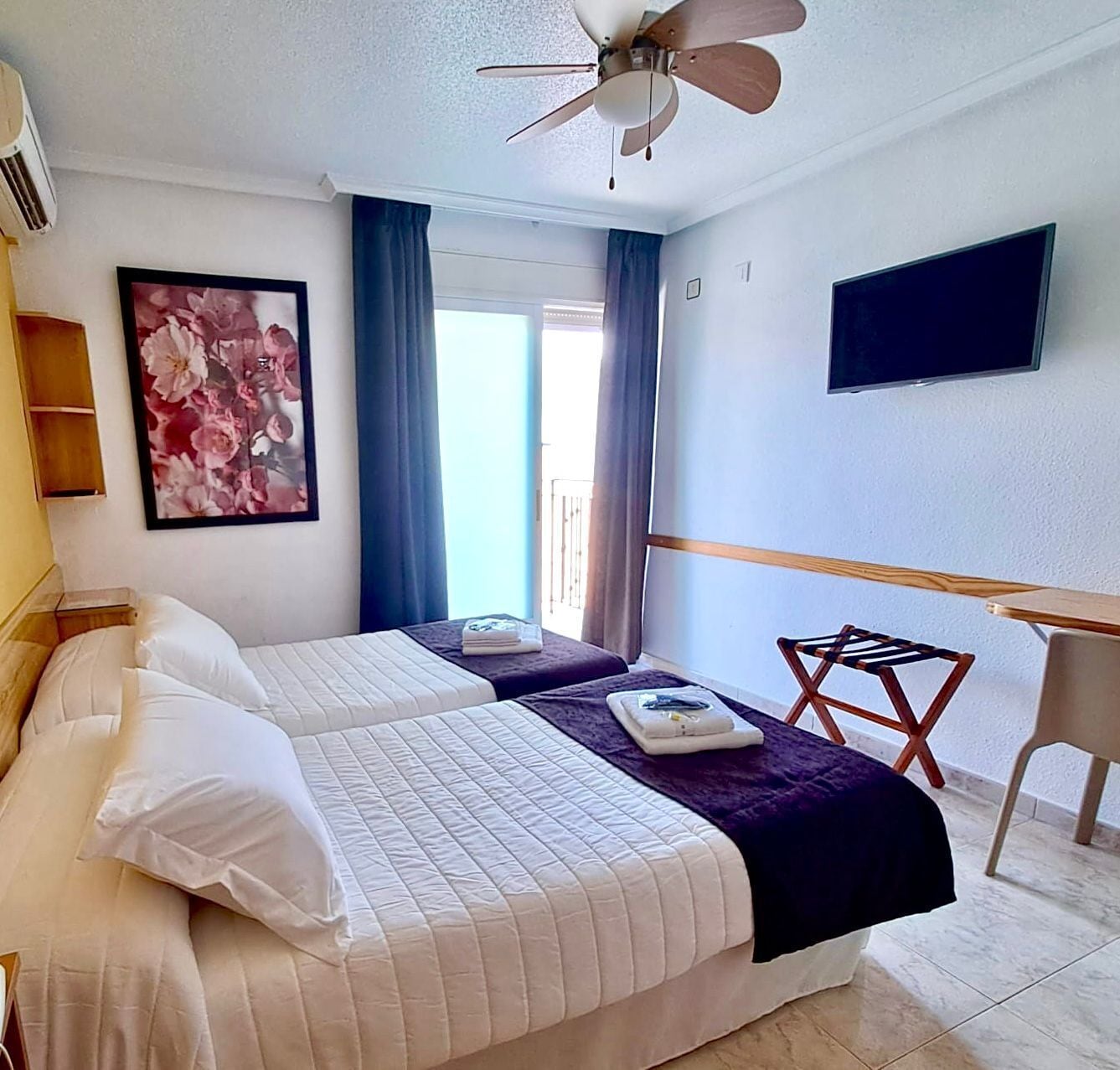 A hotel room with two beds, a ceiling fan, a TV, and a balcony.