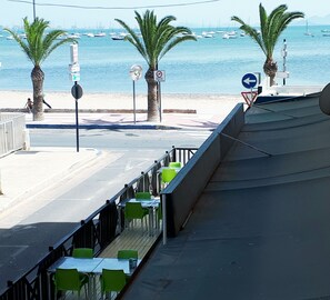Una terraza frente al mar con mesas y sillas, con vistas a una playa con palmeras y barcos en el agua.