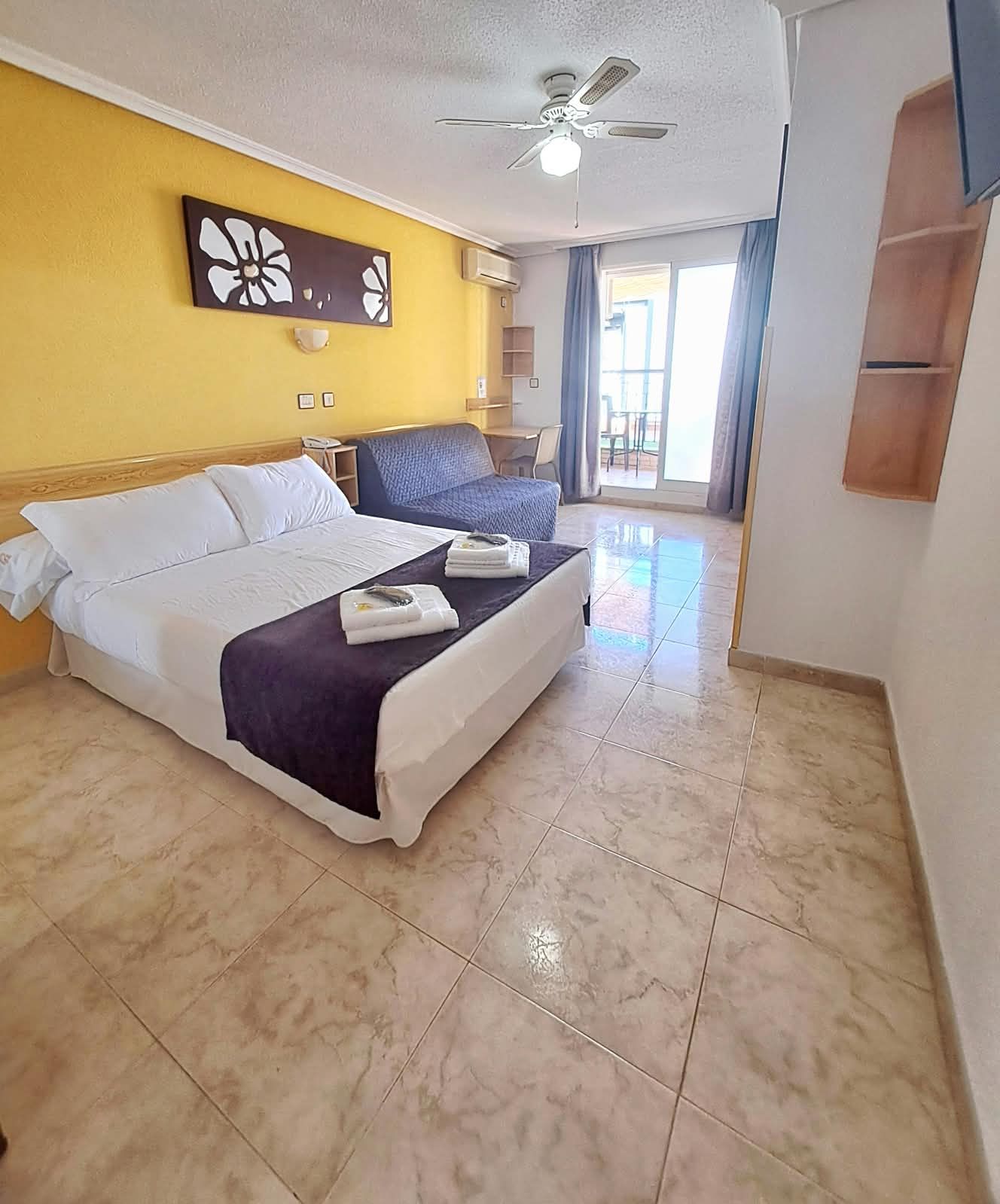 Habitación de hotel con cama, escritorio, silla, ventilador de techo y vistas al exterior.