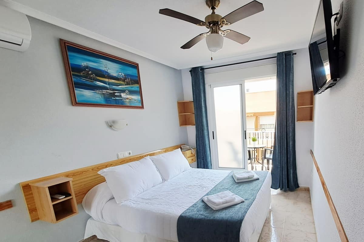Ein Hotelzimmer mit Bett, Deckenventilator, Gemälde, Fernseher und Blick ins Freie.