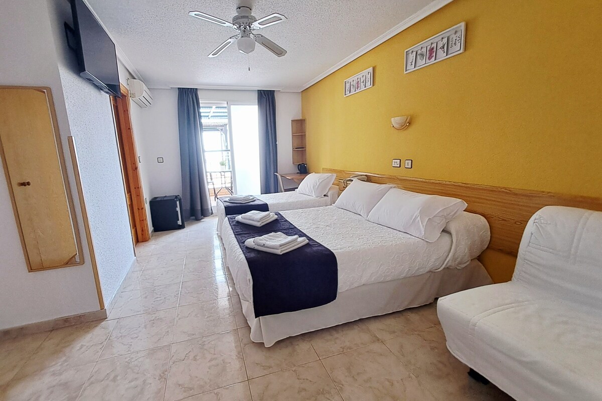 A hotel room with a bed, a sofa, a ceiling fan, a TV, and a window with curtains.